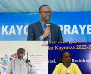 John Bosco Nyemazi speaks at a public event, standing at a podium while addressing participants during an earlier community meeting.